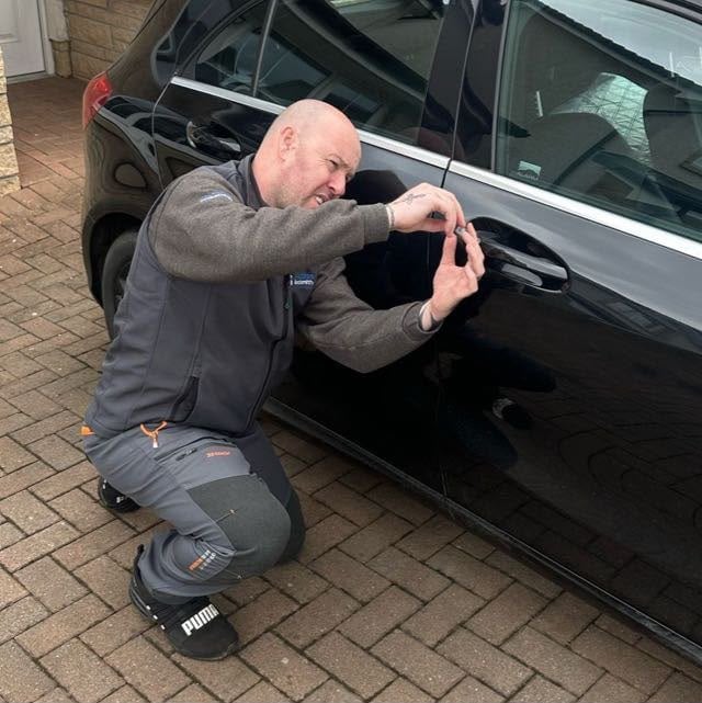 Man crouching beside a black car, examining damage to the vehicle door with his hand