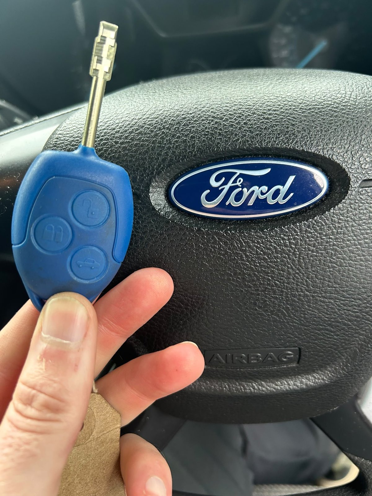 Hand holding a blue Ford car key fob in front of a steering wheel with the Ford logo